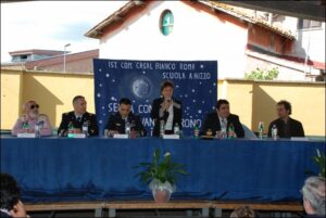 persona in piedi regge un microfono accanto a lei altre persone sedute in un banco da conferenza